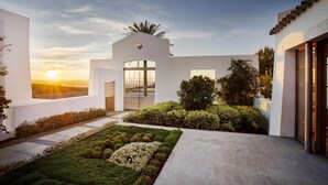 A modern building with a central entrance, a landscaped garden with various shrubs and flowers, and a view of the sunset in the background. - Exclusive Cortijo surrounded by an olive grove (Arcos de la Frontera)