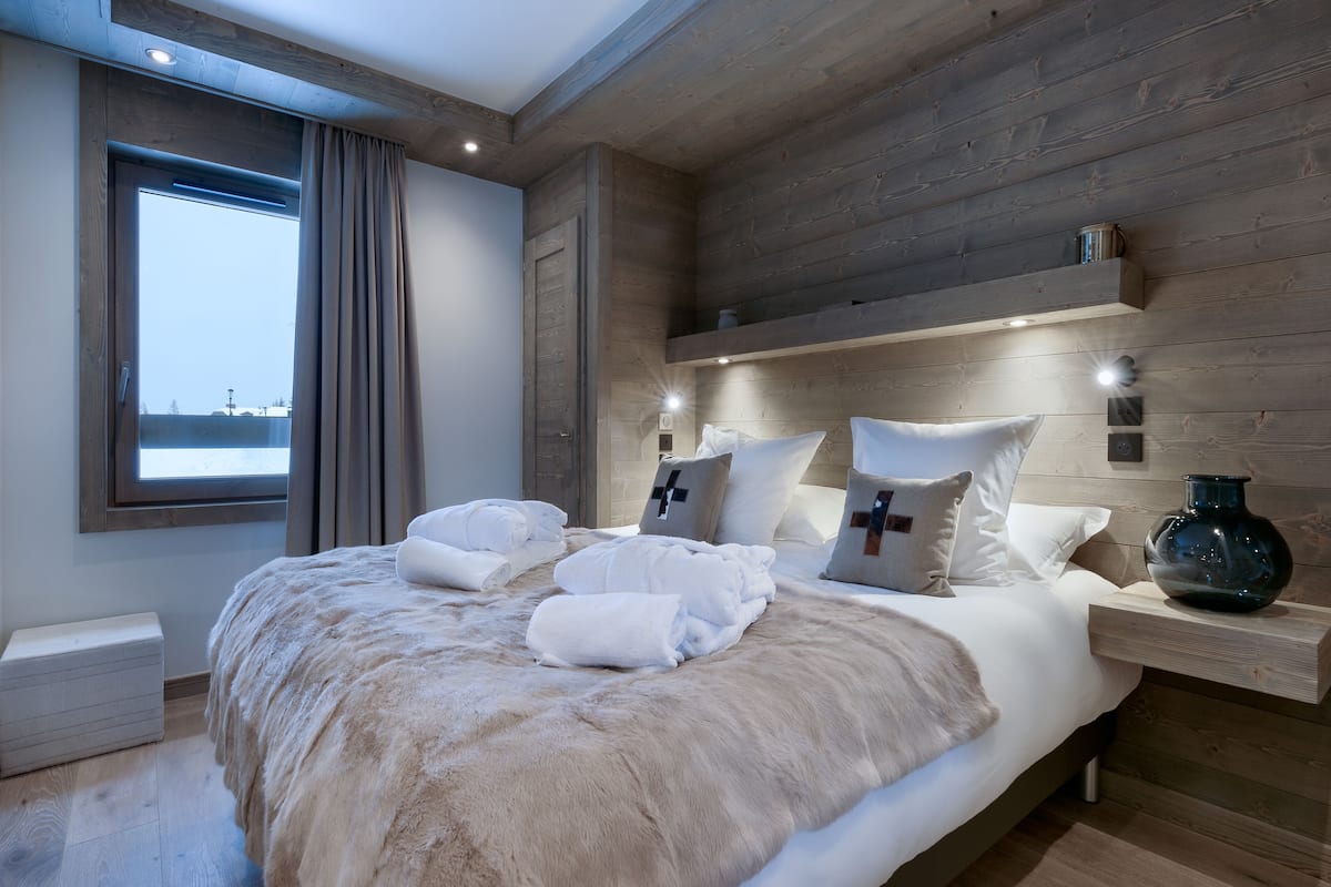 Ein modernes Schlafzimmer mit einem Holz-Kopfteil, einem großen Fenster mit Vorhängen, einem Bett mit weißen Leinen und einer Felldecke sowie einem Nachttisch mit einer Vase.