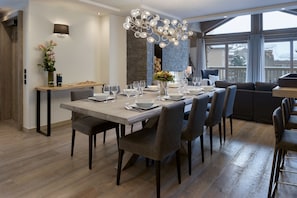 Un comedor moderno con una gran mesa de madera dispuesta para seis personas, rodeada de sillas tapizadas en gris. Un candelabro único cuelga sobre la mesa, y un jarrón con flores está colocado en el lado izquierdo de la mesa.