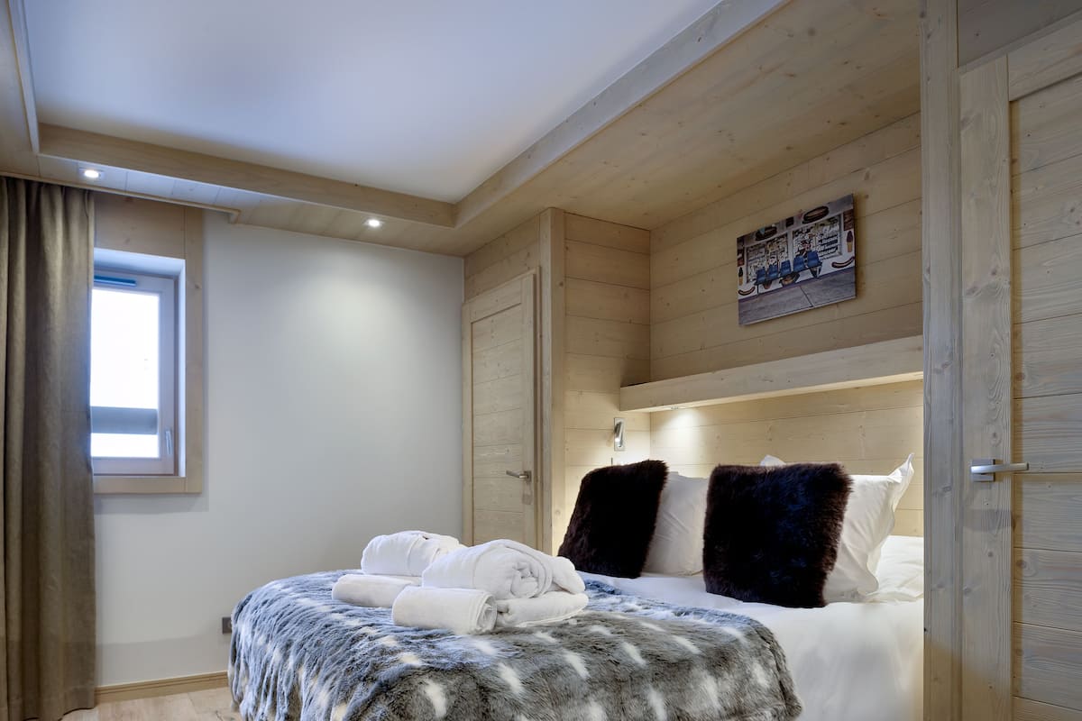 Ein Schlafzimmer mit Holzbalkendecke, einem Bett mit weißen Leinen und einem Fellüberwurf, einem Fenster mit Vorhängen und einem Wandbild.