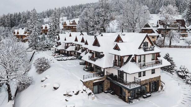 Um chalé coberto de neve com vários edifícios no estilo de chalé, um edifício central com varanda e uma floresta ao redor.