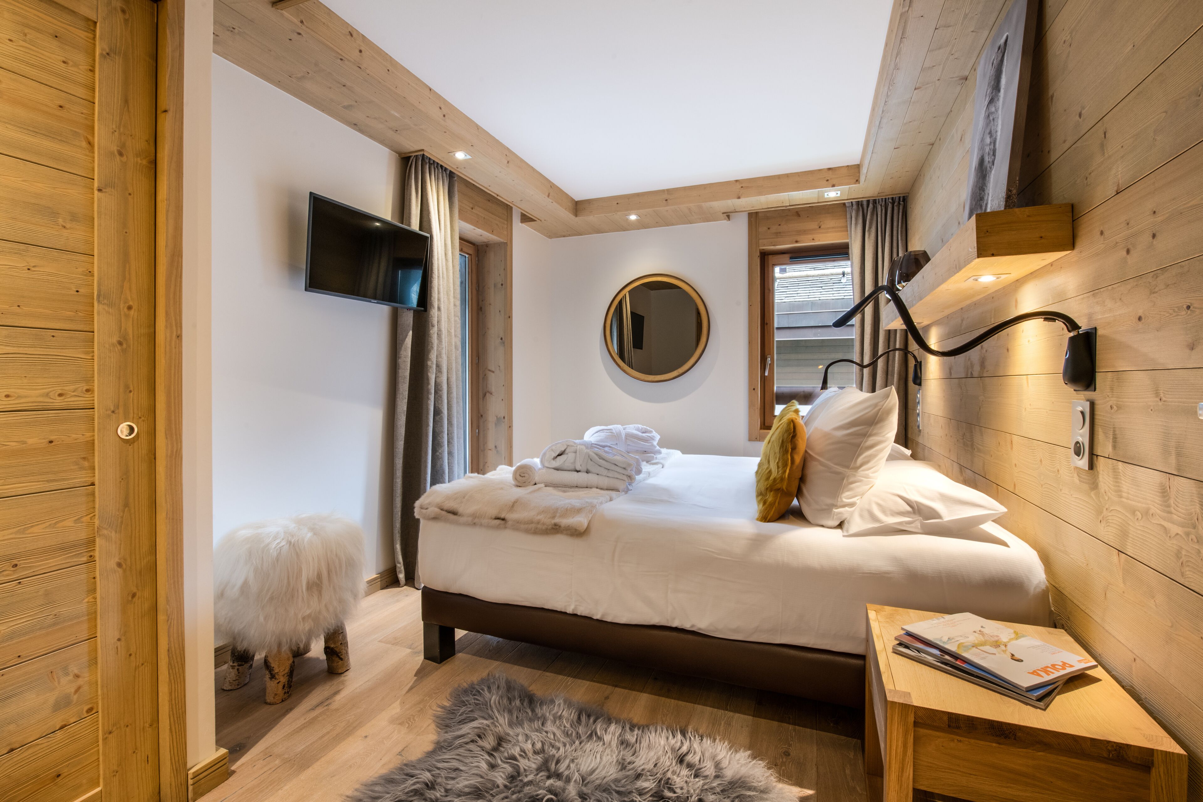 A bedroom with a wooden ceiling, a bed with white linens, a fluffy rug, and a small wooden nightstand with a magazine.