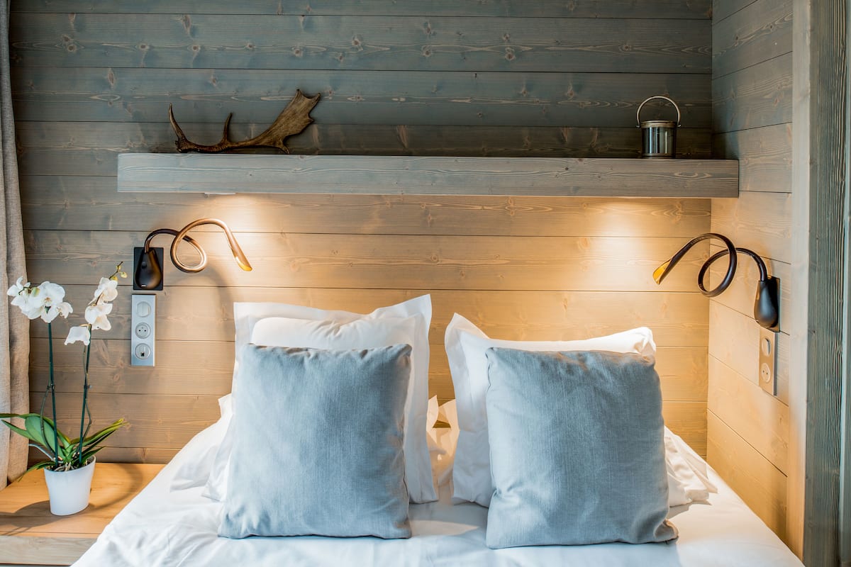 Ein holzgetäfeltes Zimmer im Blockhausstil mit einem Bett, Kissen, einem Nachttisch, einer Pflanze und Wandleuchten.
