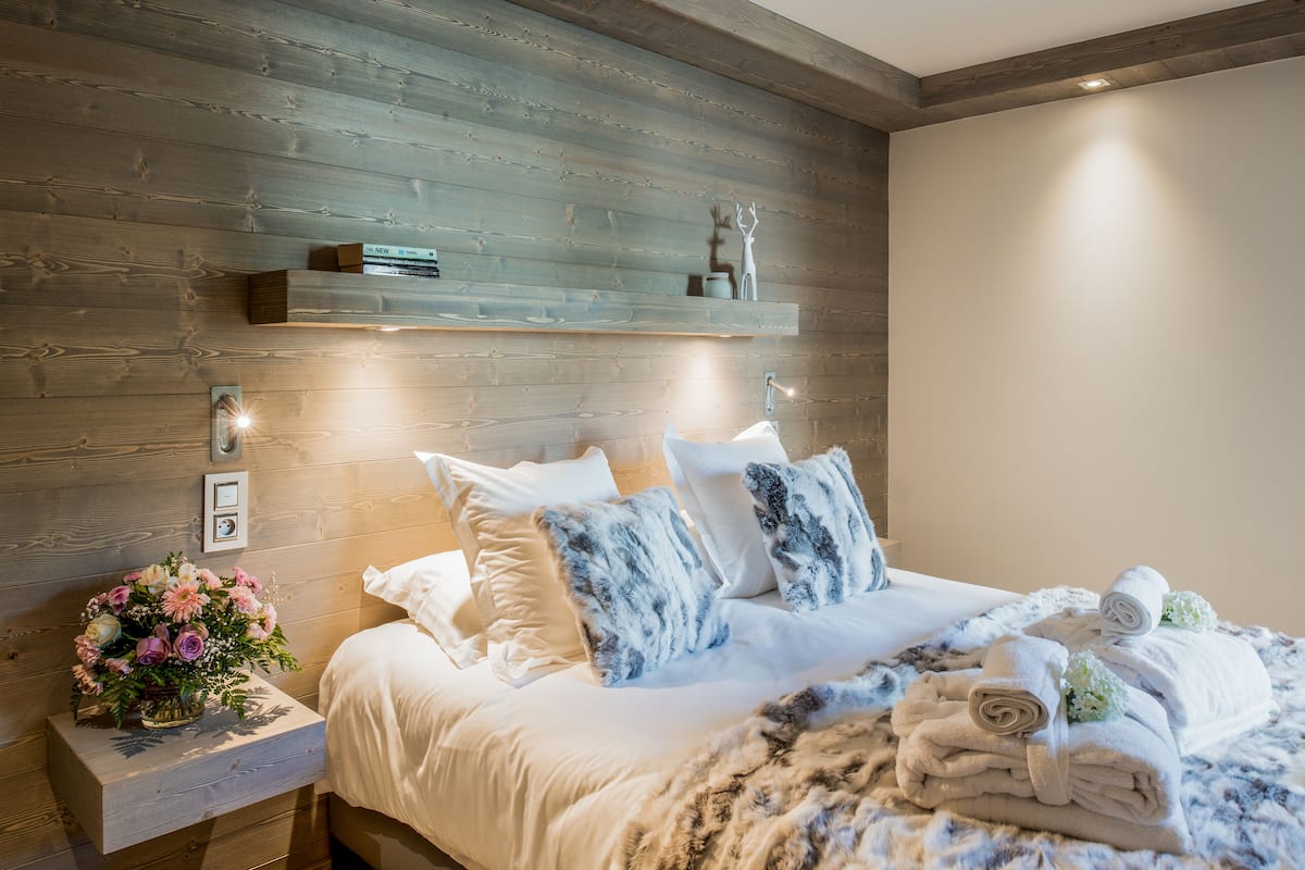 Ein ordentlich bezogenes Bett mit weißen Leinen und Kissen in Blau-Weiß, ein Holz-Kopfschild und ein Nachttisch mit einem Blumenstrauß.
