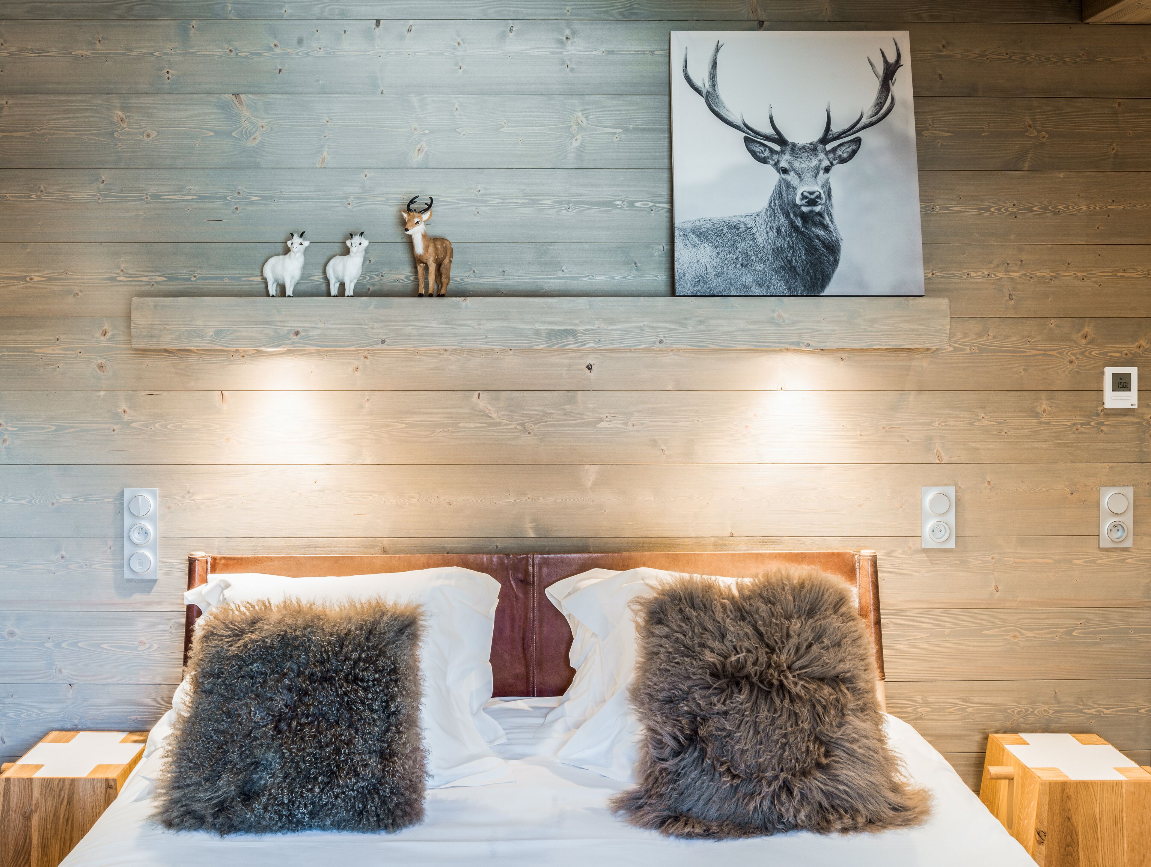 Une chambre avec un mur en bois, un lit avec deux oreillers moelleux, un tableau représentant un cerf et un thermostat fixé au mur.
