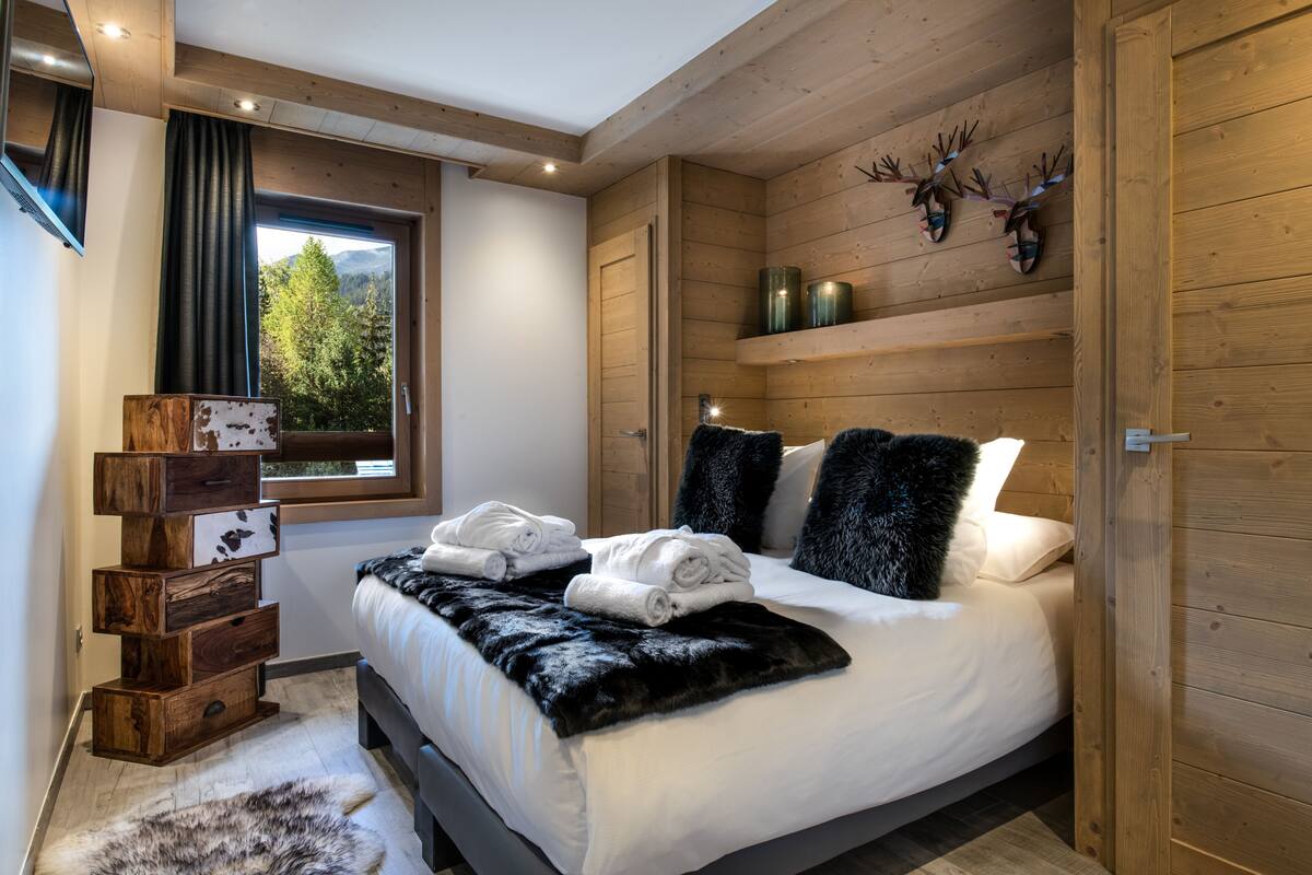Ein Schlafzimmer mit Bett, einem fensterförmigen Ausblick, Holzwänden, einer hölzernen Kommode und einem Fellüberwurf.