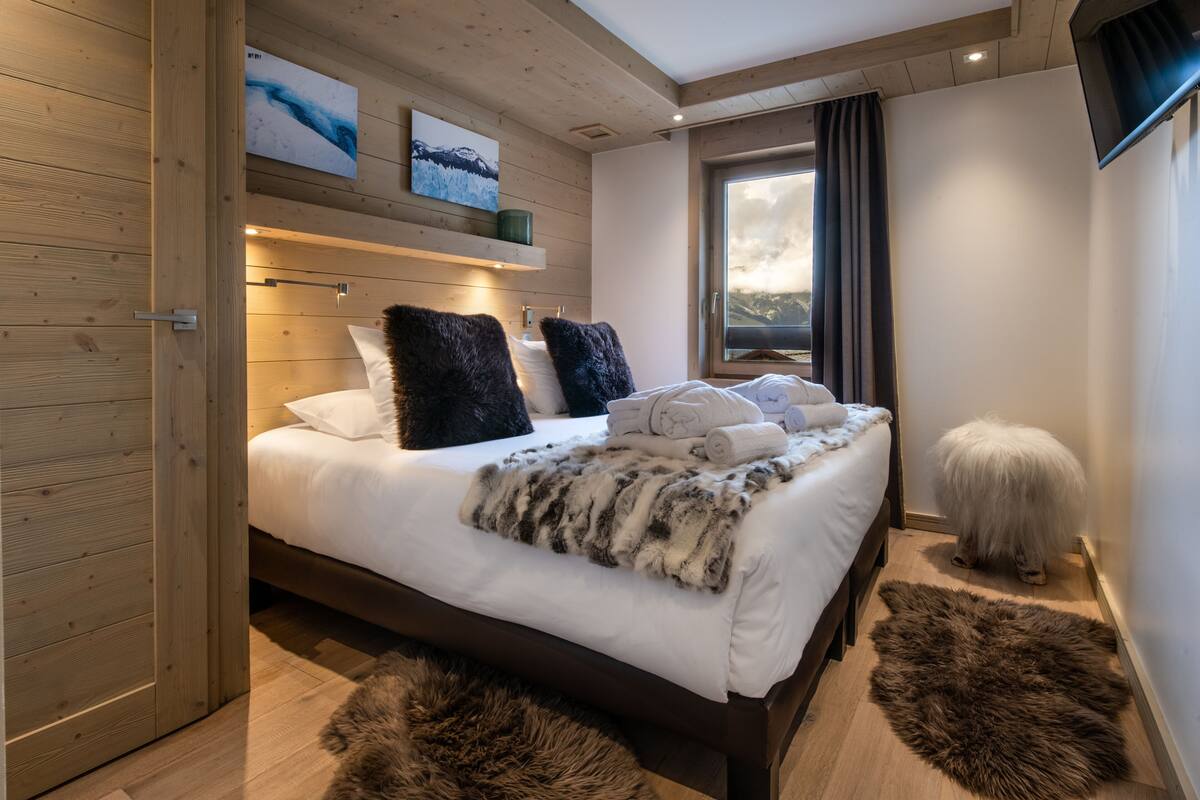Ein modernes Schlafzimmer mit Holzboden, einem großen Bett mit flauschigen Kissen, einer Felldecke und Blick auf Berge durch ein Fenster.