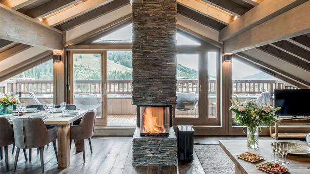 Una habitación acogedora con chimenea de piedra, vigas de madera, zona de comedor y grandes ventanales con vistas a la montaña.
