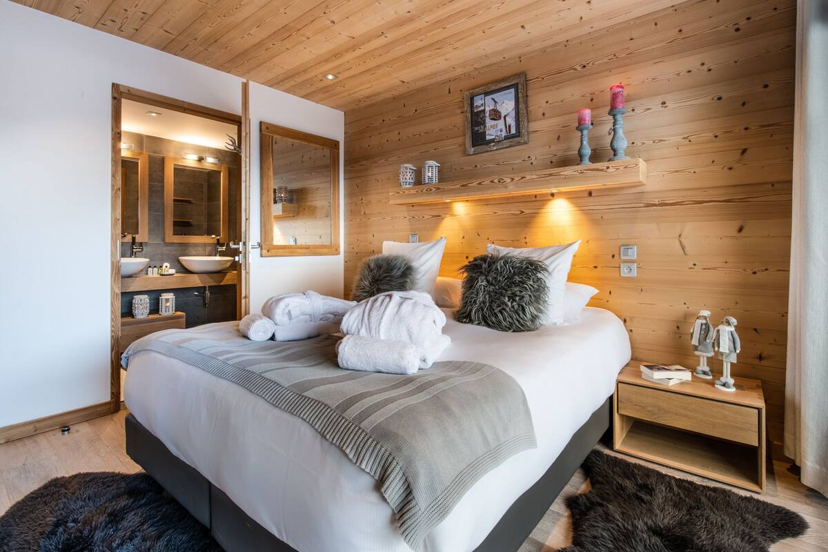 Un dormitorio moderno con techo de madera, una cama grande con manta a rayas gris y blanco, una mesita de noche con lámpara y una mesita auxiliar de madera con objetos decorativos.