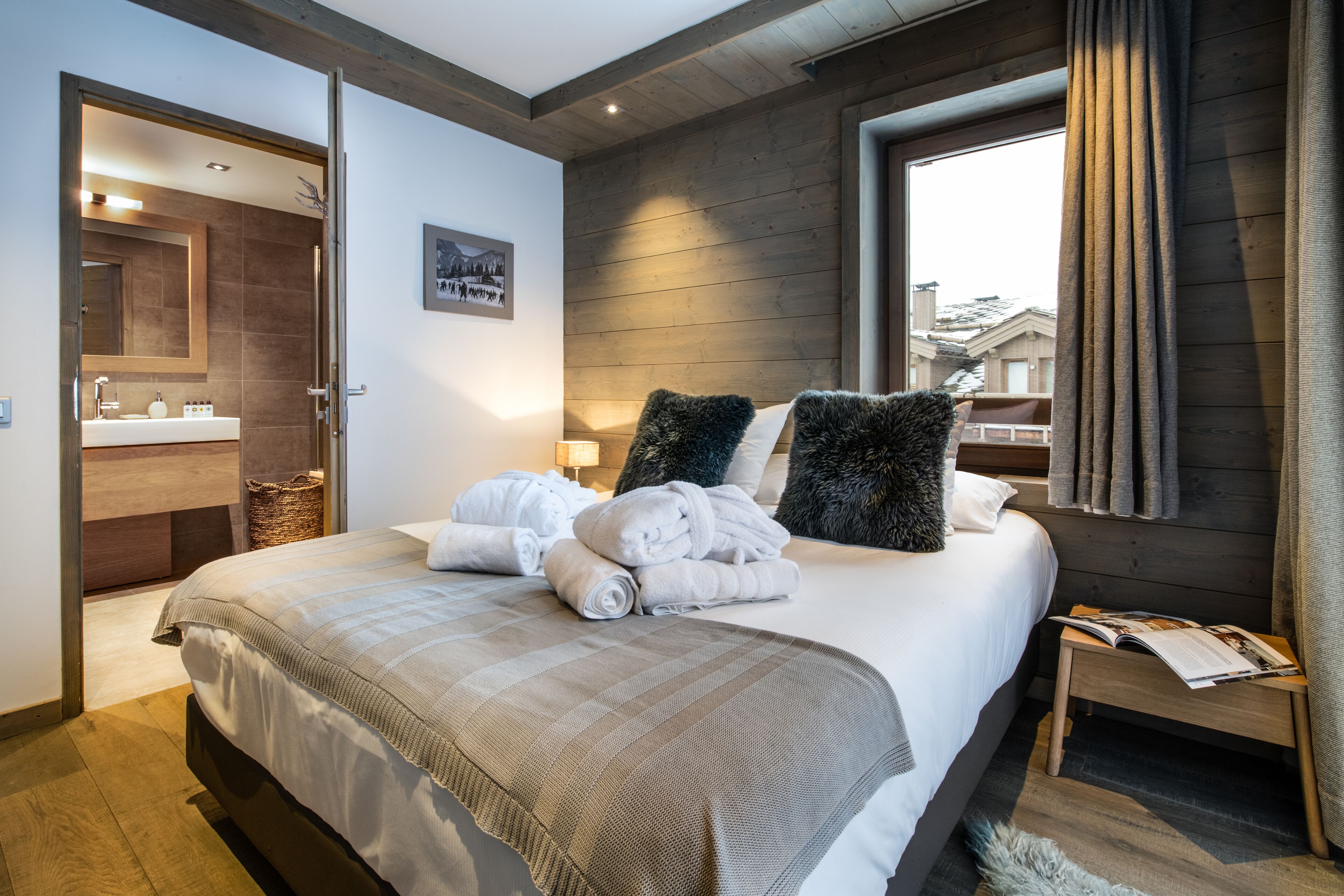 A modern bedroom with a large bed, a wooden headboard, a bedside table with a book, and a view of the outside through a window.