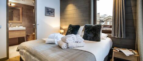A modern bedroom with a large bed, a wooden headboard, a bedside table with a book, and a view of the outside through a window.