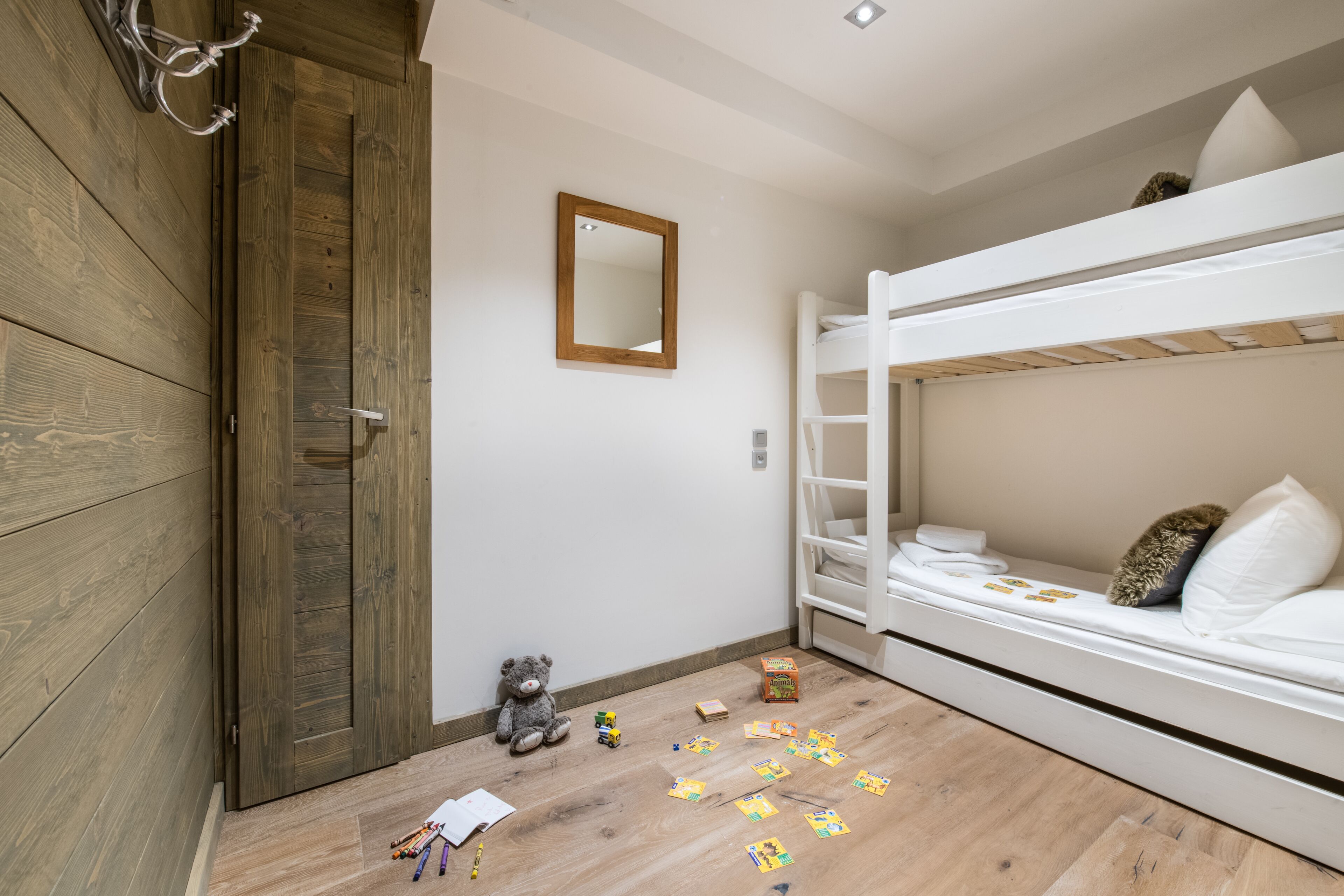 A child's room with a bunk bed, wooden flooring, a teddy bear, and scattered toys.