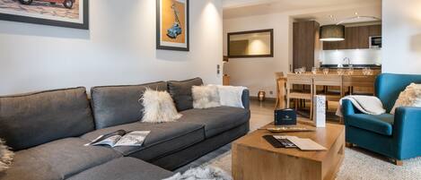 Una sala moderna con un sofá gris, un sillón azul y una mesa de centro de madera. Se observa una cocina al fondo con armarios de madera y una mesa de comedor.