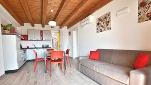 Ein modernes Wohnzimmer mit grauem Sofa und roten Sesseln, einer Holzbalkendecke sowie einer Küche mit weißen Schränken und roten Akzenten.