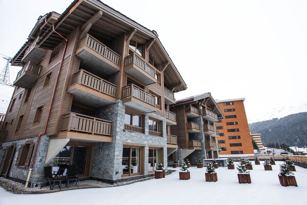 Un hotel de varios pisos con balcones, fachada de piedra y el paisaje de una estación de esquí al fondo.
