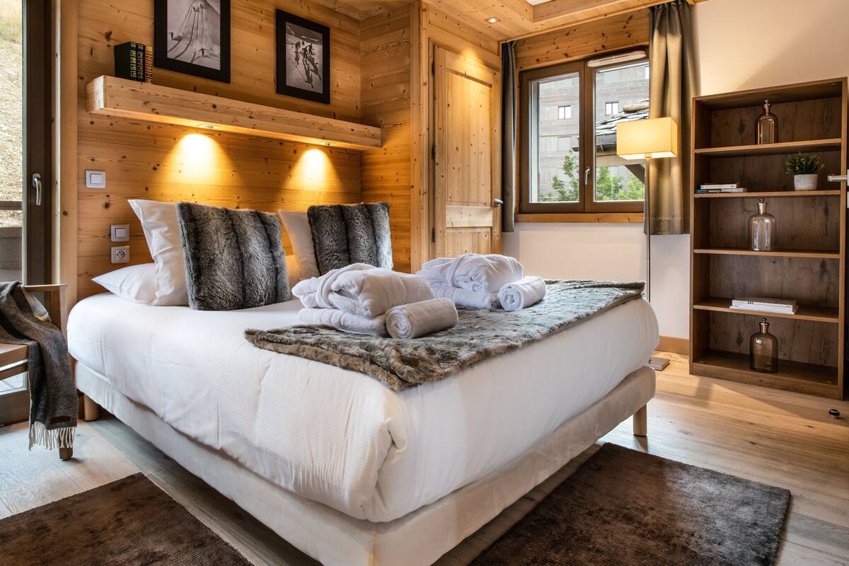 A wooden bedroom with a large bed, two bedside lamps, a wooden shelf with books, and a window with curtains.