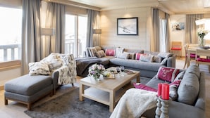 A modern living room with a grey sofa, ottoman, and coffee table, a large window with curtains, and a painting on the wall. - Aspen Lodge by Alpine resorts (Courchevel)