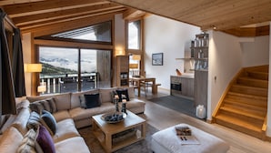 A spacious living area with a large sofa, a coffee table, and a view of the mountains through large windows. There is a wooden staircase leading to an upper level. - Aspen Lodge by Alpine resorts (Courchevel)