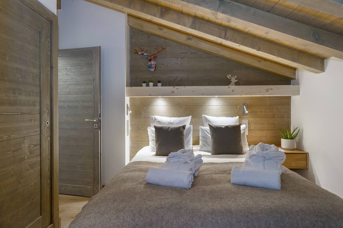 A bedroom with a wooden ceiling, a bed with white bedding, two bedside tables with potted plants, and a wall-mounted deer head decoration.