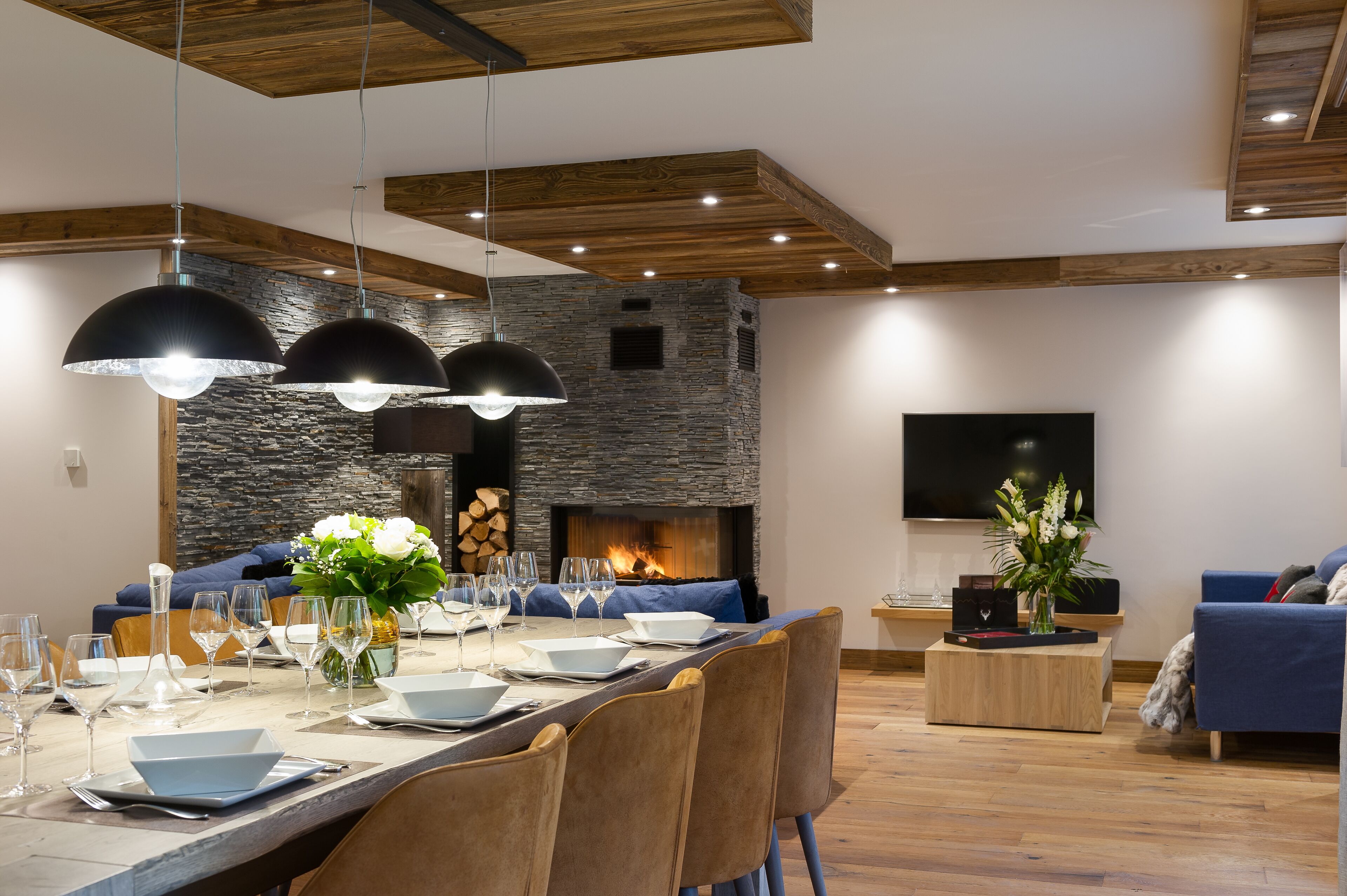 A modern dining area with a stone fireplace, wooden ceiling beams, and a long dining table set for six.