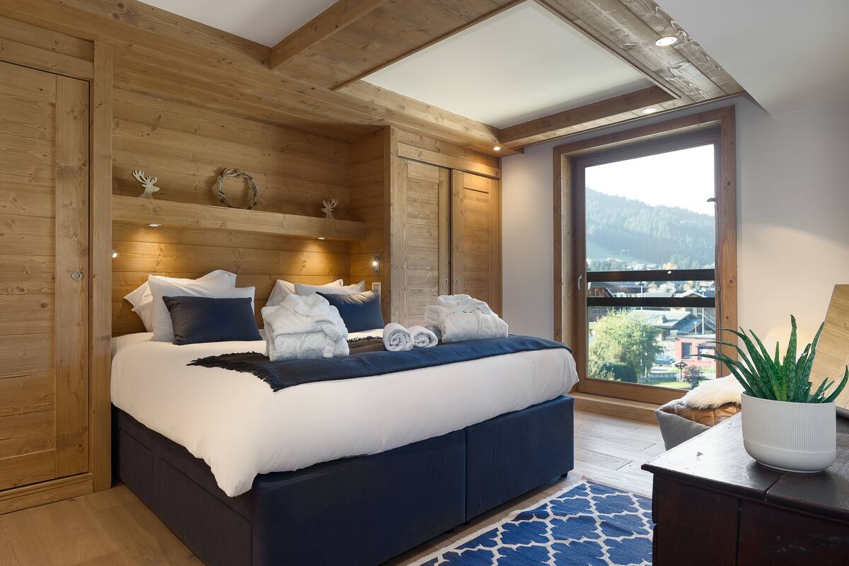 A bedroom with a wooden ceiling, a large window with a view, a bed with white and blue bedding, and a potted plant on a dresser.