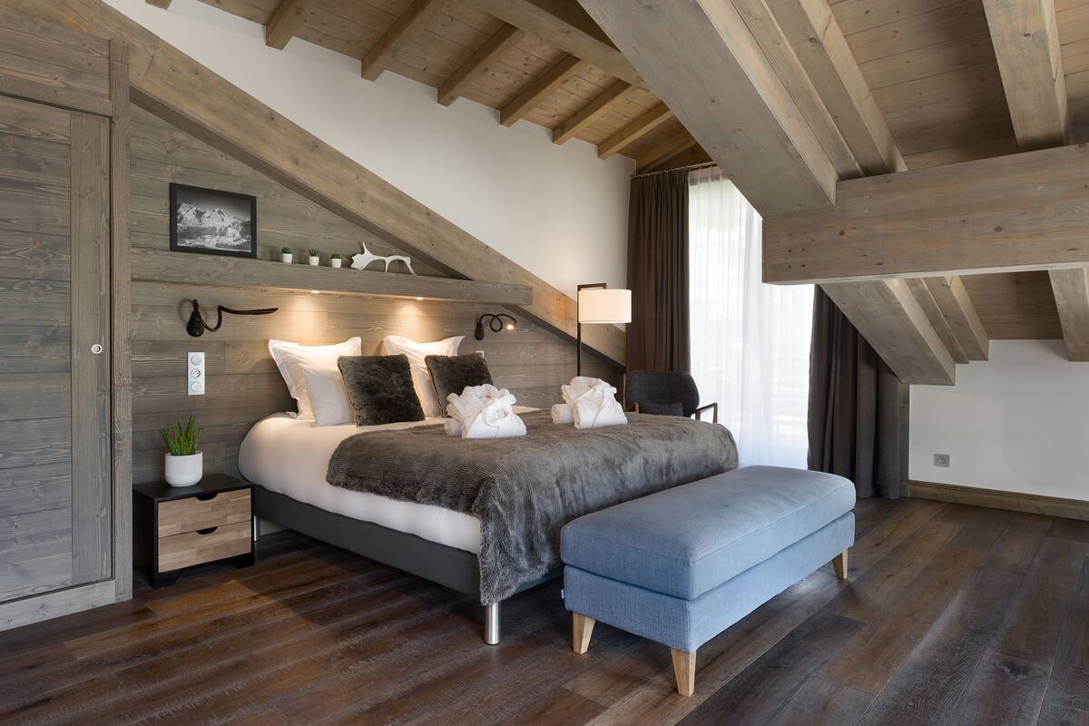 A bedroom with a sloped ceiling, wooden beams, a bed with pillows and towels, a bedside table with a plant, and a lamp.