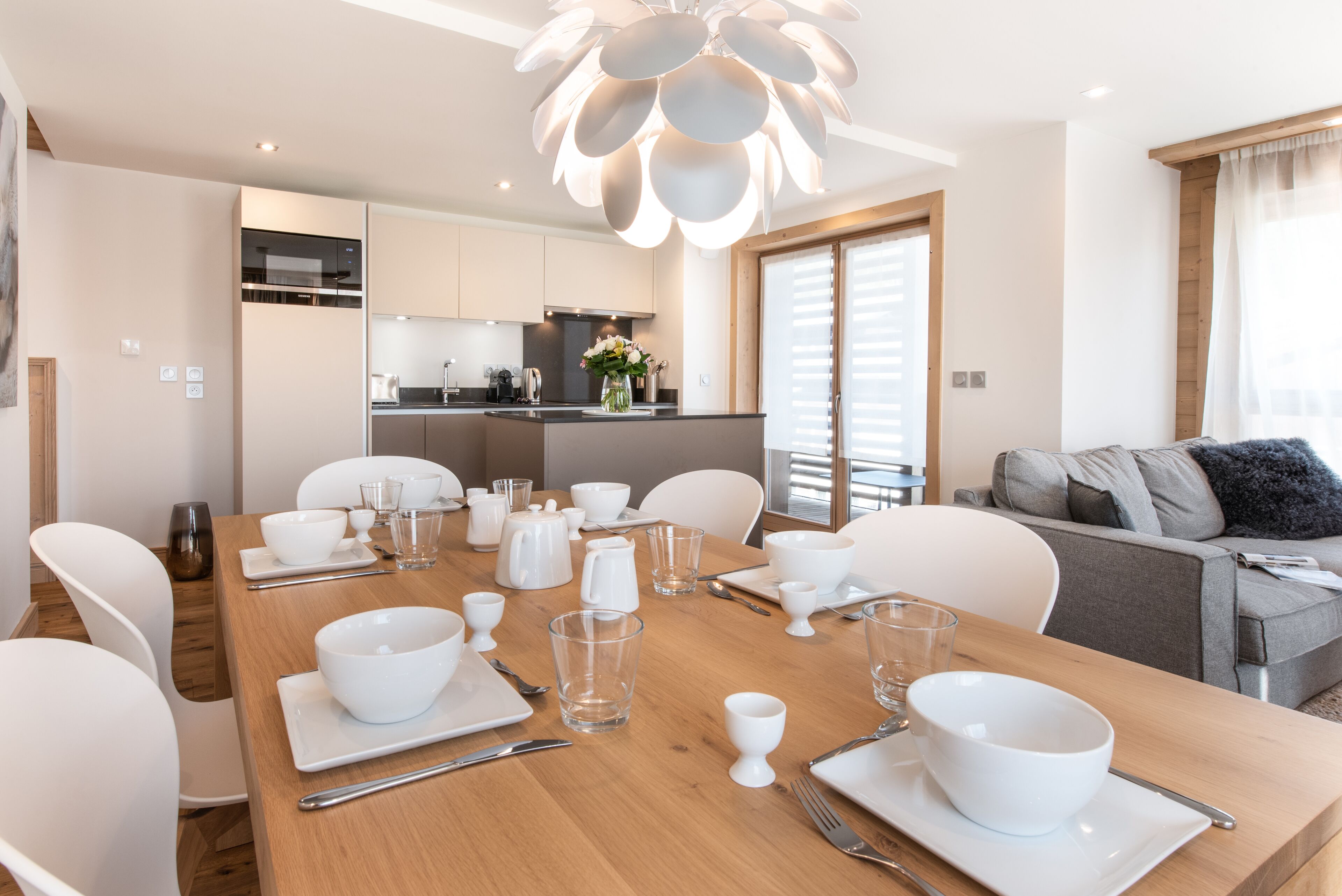 Un comedor moderno con una mesa de madera dispuesta para seis comensales, sillas blancas y una cocina al fondo.