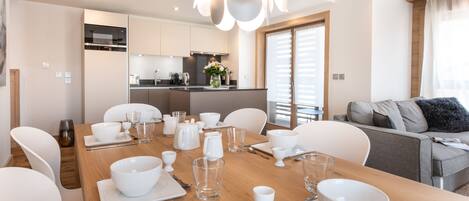 Un comedor moderno con una mesa de madera dispuesta para seis comensales, sillas blancas y una cocina al fondo.