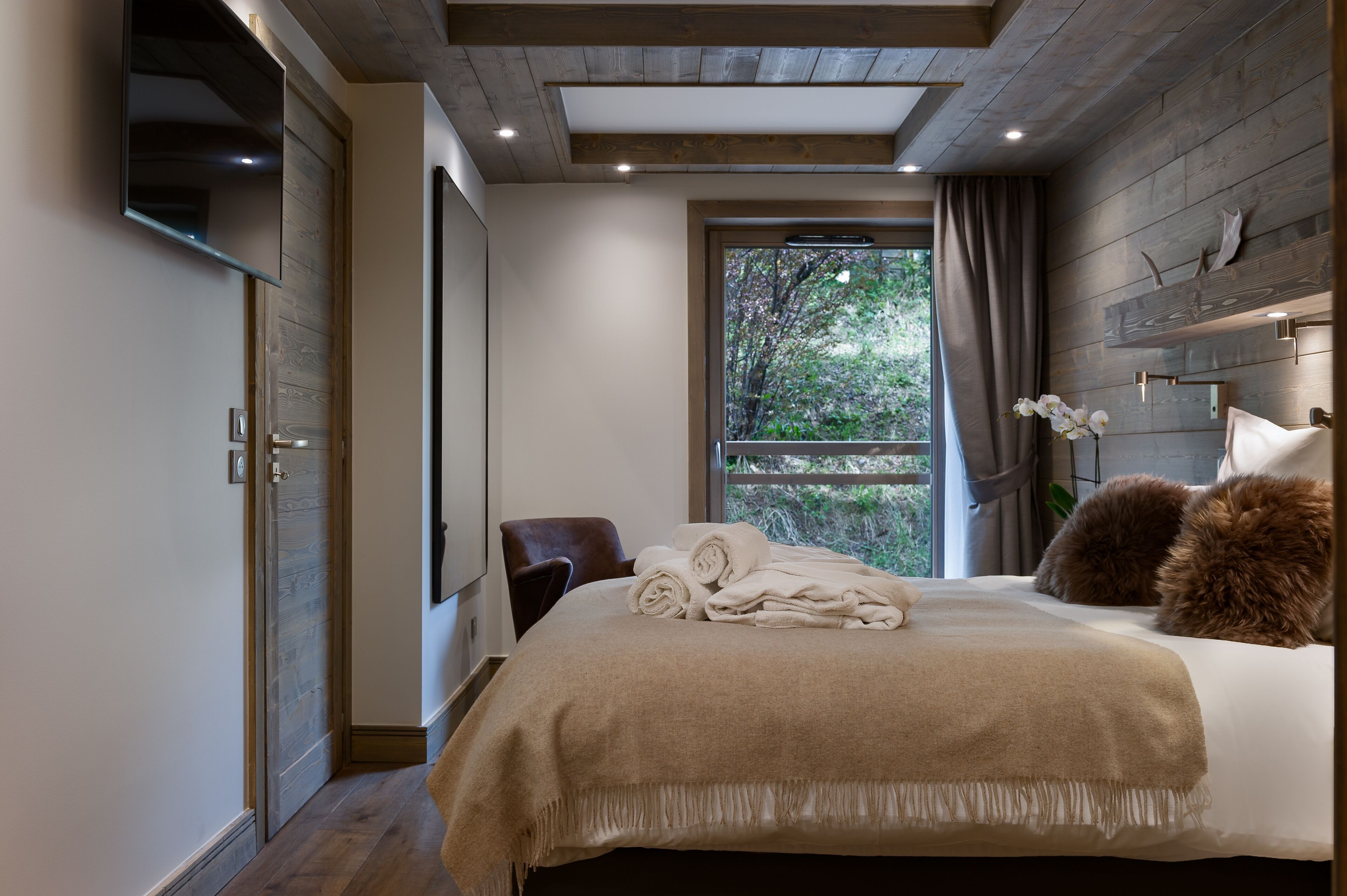 Une chambre à coucher moderne avec un grand lit, une tête de lit en bois, une fenêtre donnant sur les arbres et un téléviseur à écran plat fixé au mur.