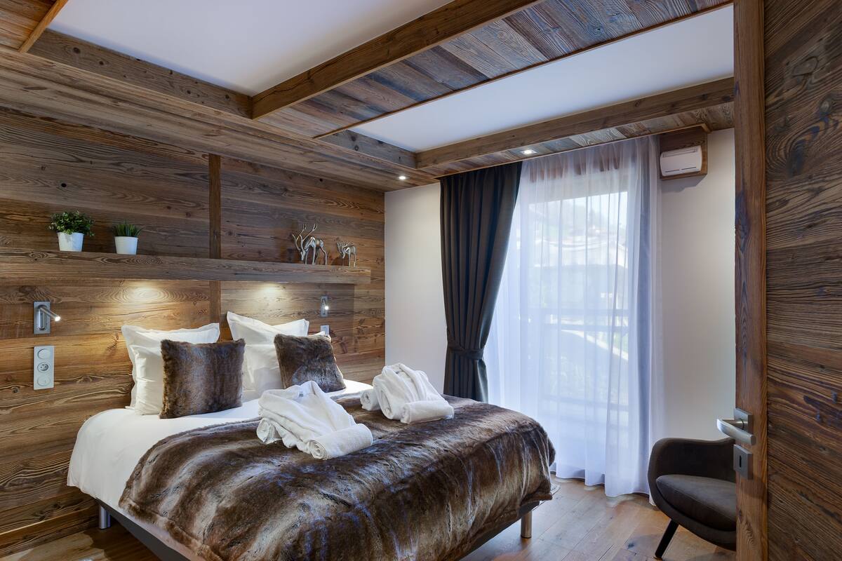 A modern bedroom with a wooden ceiling, a large bed with white linens, a fur throw, and a chair.