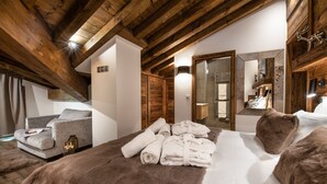 A cozy bedroom with a wooden ceiling, a bed with a fur throw, a grey sofa, and a bathroom visible through an open door.