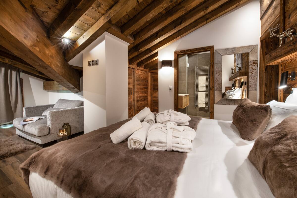 A cozy bedroom with a wooden ceiling, a bed with a fur throw, a grey sofa, and a bathroom visible through an open door.