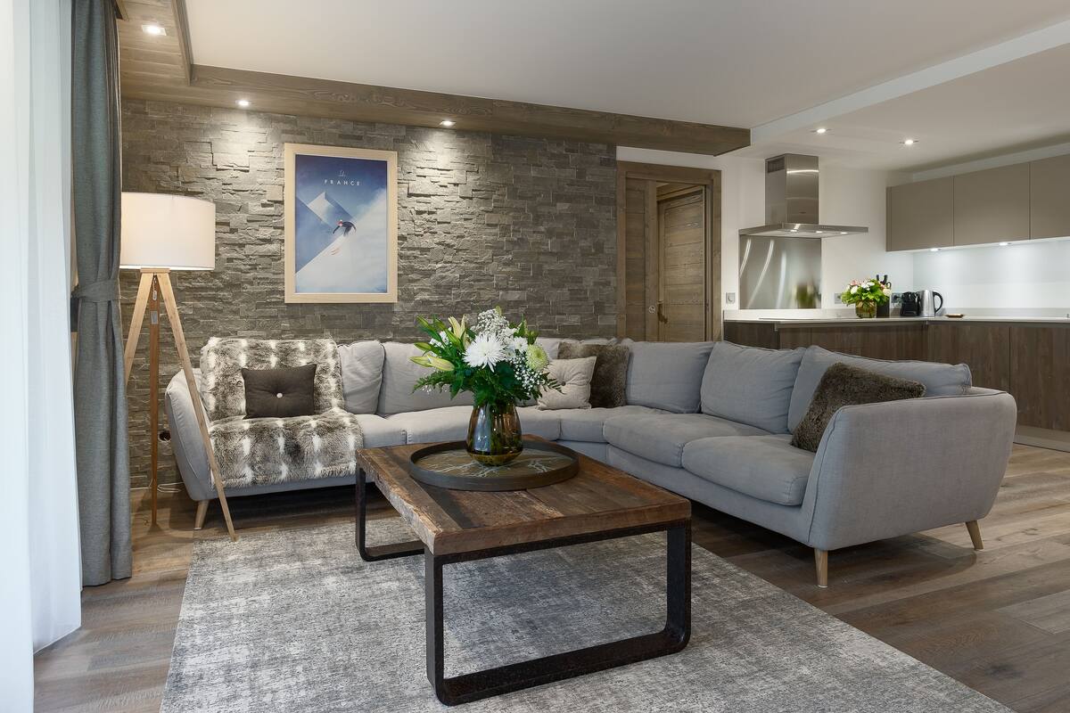 A modern living room with a grey sofa, a wooden coffee table with a vase of flowers, and a stone wall.