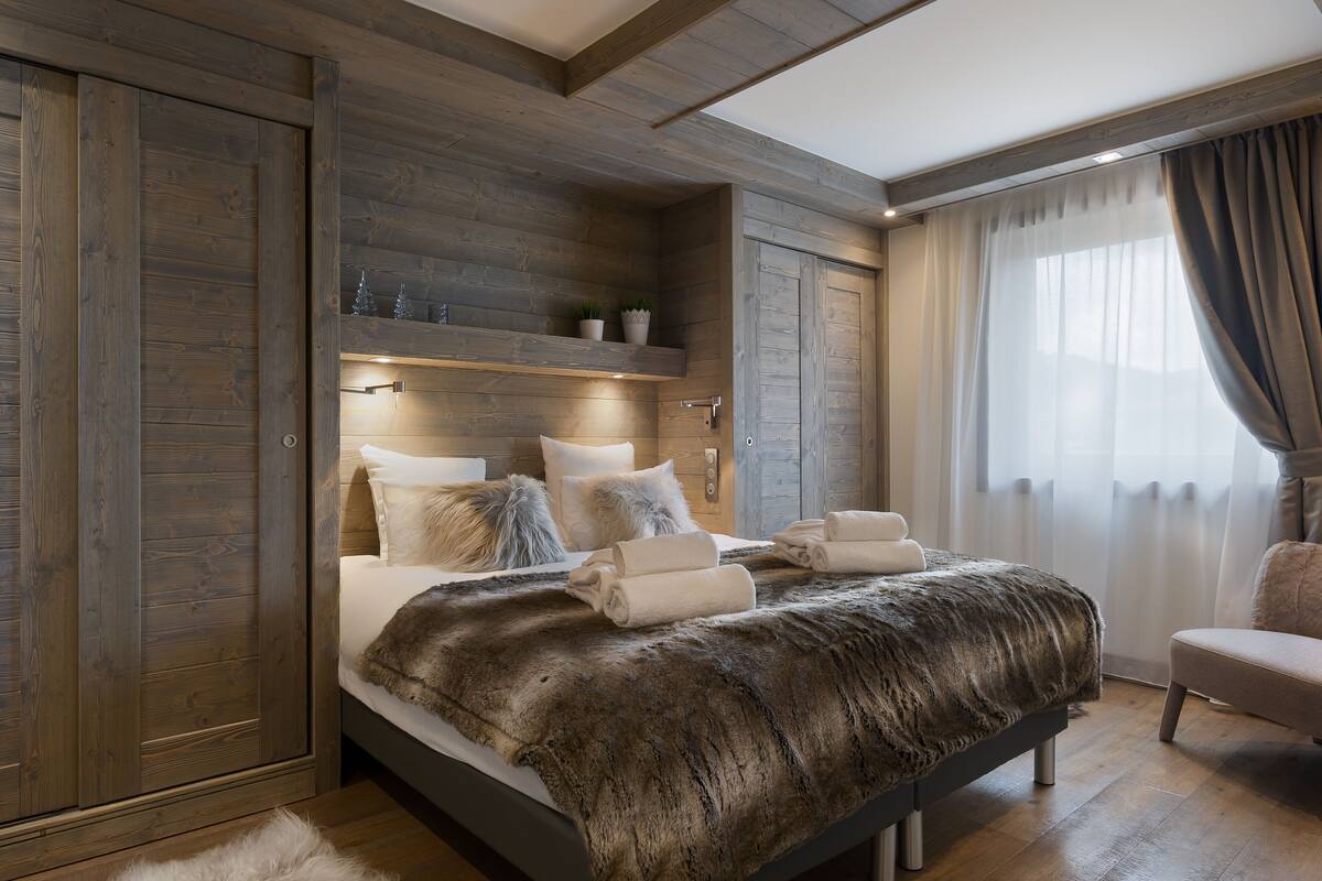 A bedroom with a large bed, wooden paneling, a built-in shelf with decorative items, and a view of the outdoors through a window with curtains.