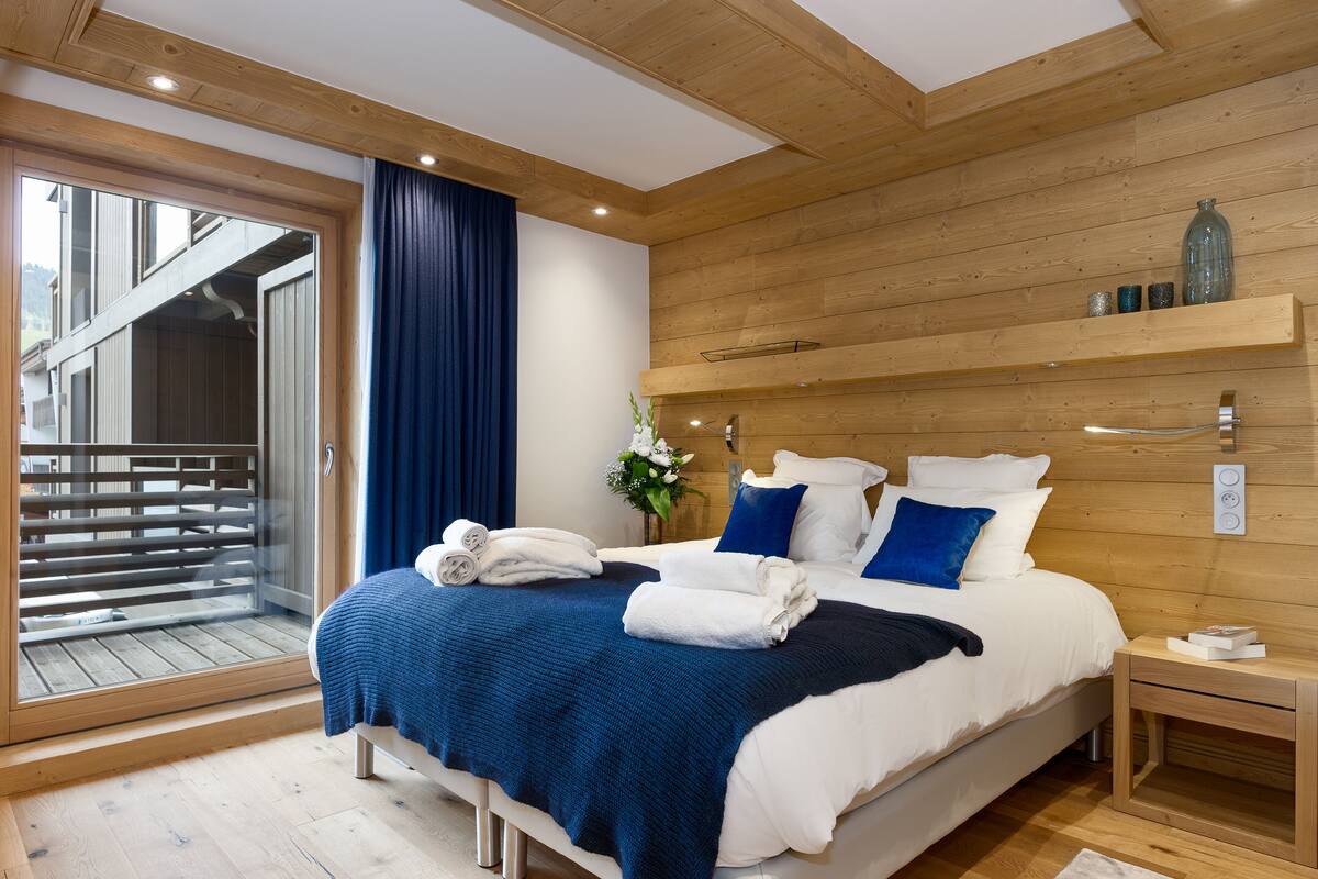 A modern bedroom with a large bed, wooden walls, a balcony, and a small bedside table.