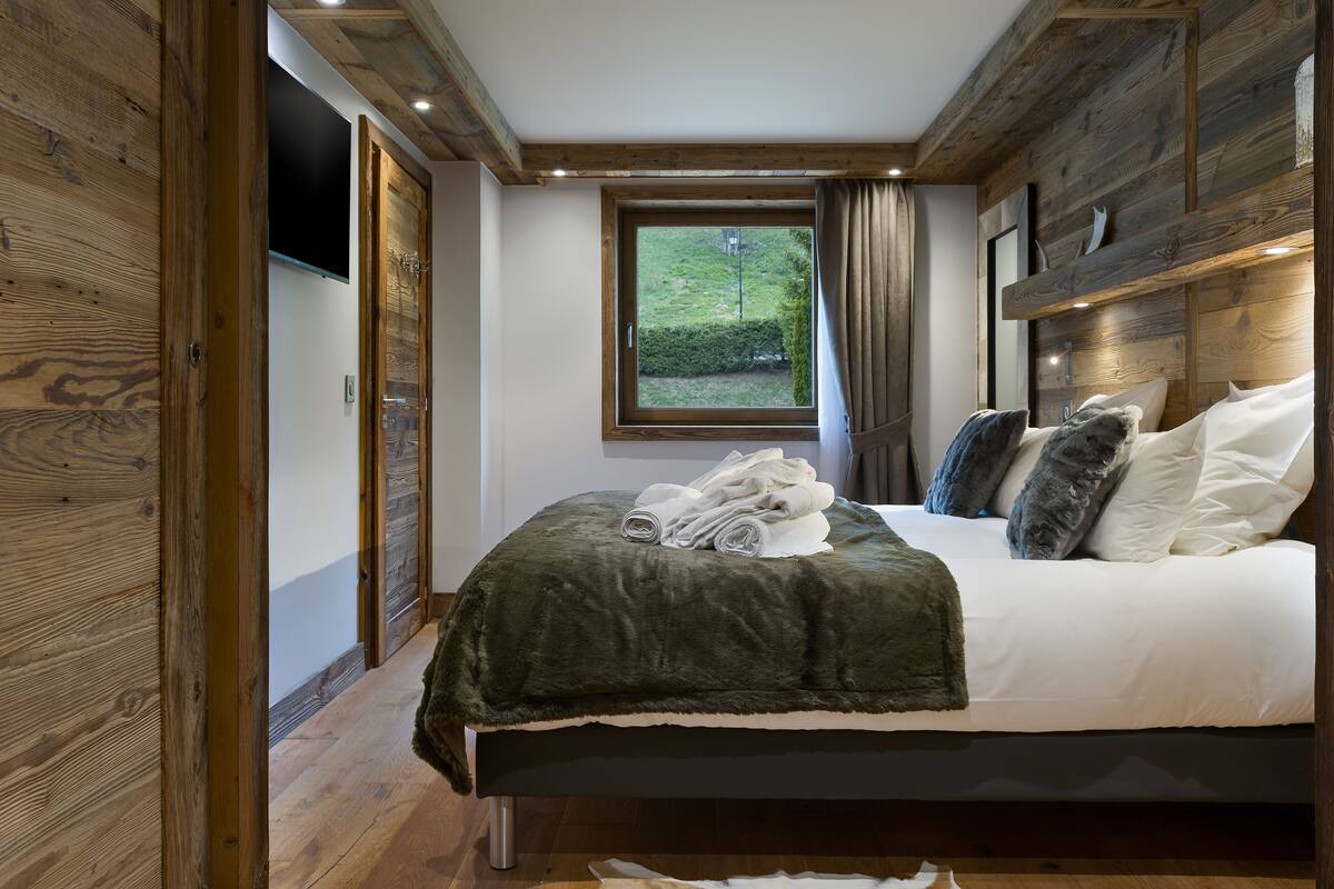 A bedroom with a wooden ceiling, a large window, a bed with white linens, and a dark green blanket.