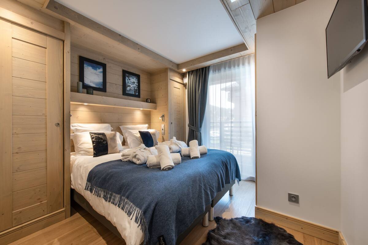A modern bedroom with a large bed, wooden paneling, a mounted TV, and a view of the outdoors.