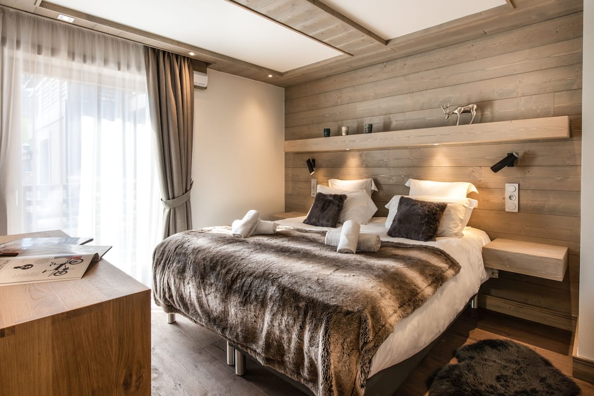 A modern bedroom with a wooden headboard, a large bed with a fur throw, bedside tables, and a desk with a lamp.