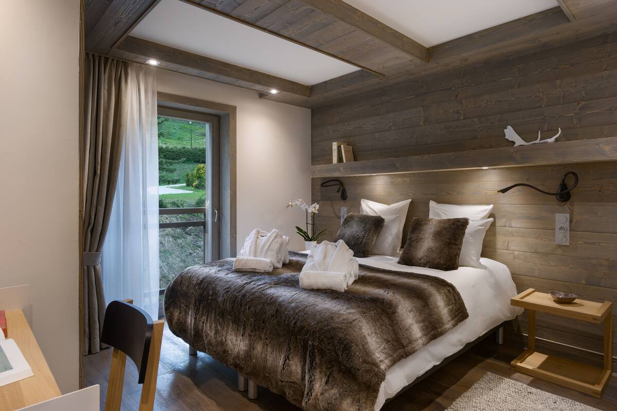 A modern bedroom with a large bed, wooden headboard, and a view of greenery outside.