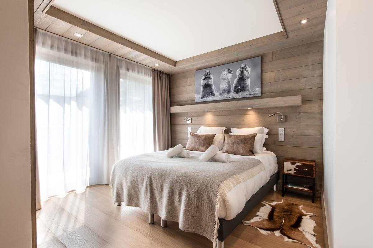 A modern bedroom with a wooden headboard, a large bed with white bedding, a mounted artwork above the bed, and a small side table with a leather item.