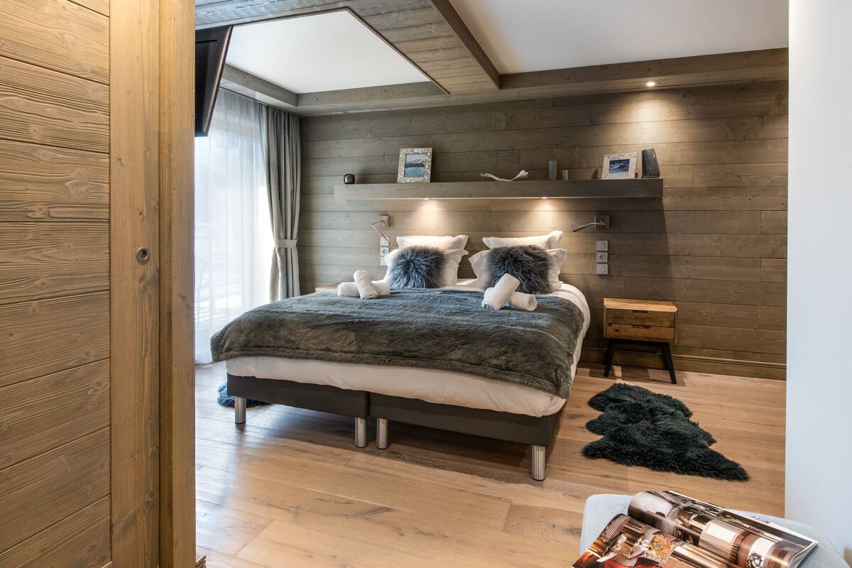 A modern bedroom with a wooden floor, a bed with a dark blanket and pillows, a nightstand, and a shelf with decorative items.