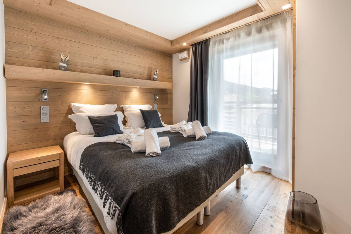 A modern bedroom with a wooden headboard, a bed with white and dark pillows, a fur rug, and a view of a balcony through sheer curtains.