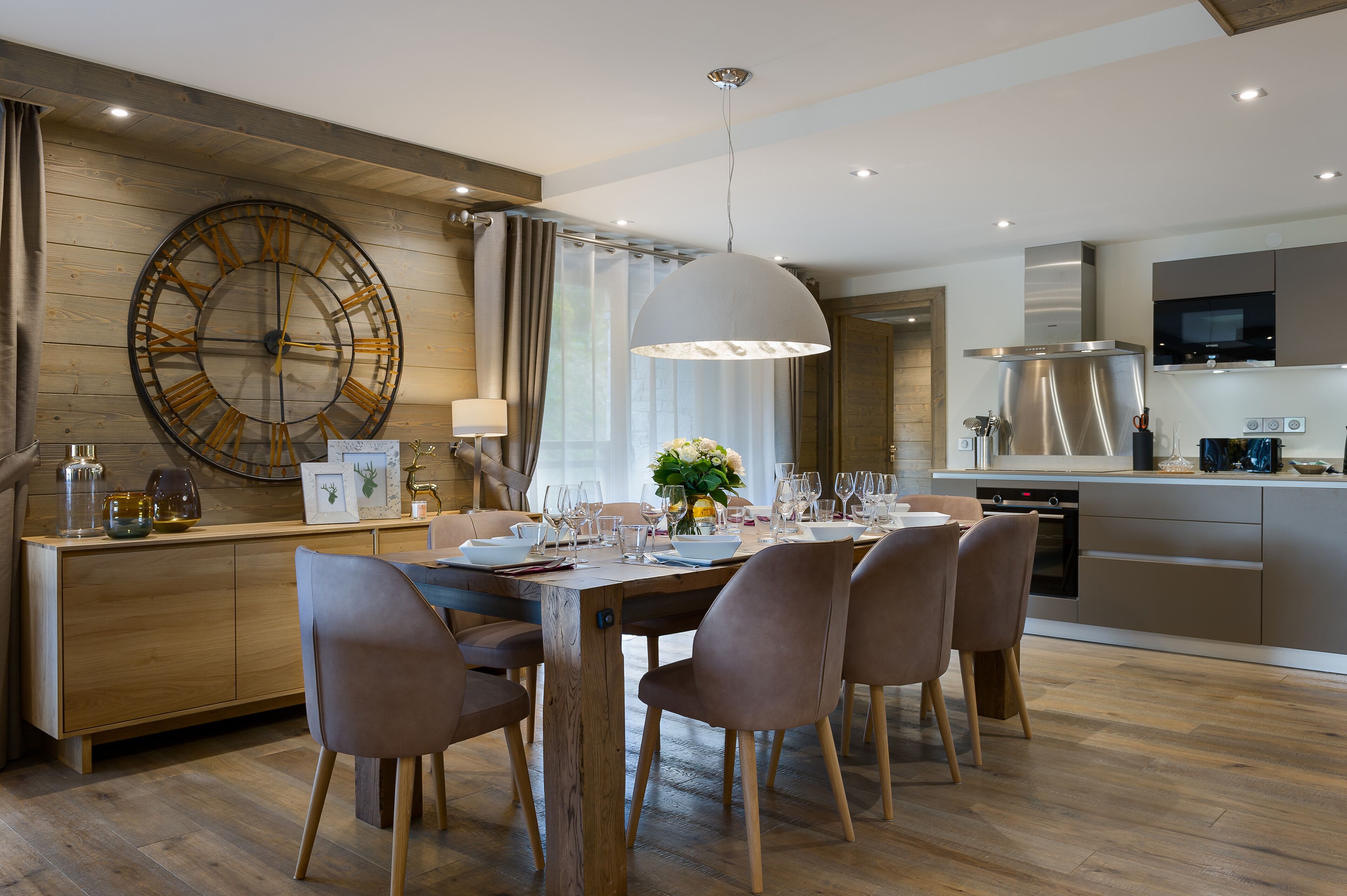 A modern kitchen with a large clock, wooden cabinetry, and a dining area with a table set for four.
