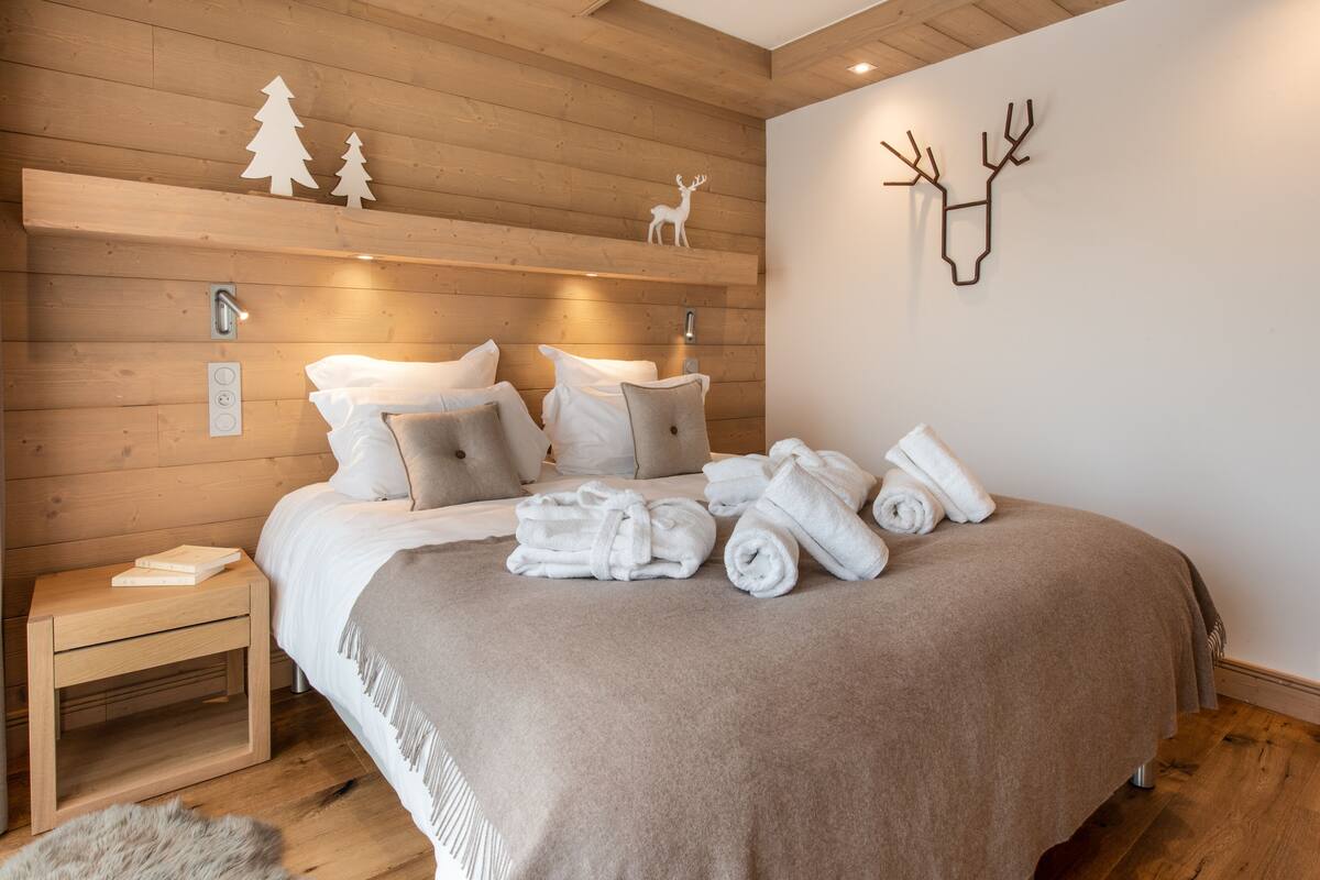 A neatly made bed with a brown blanket, white pillows, and rolled towels on a wooden floor.