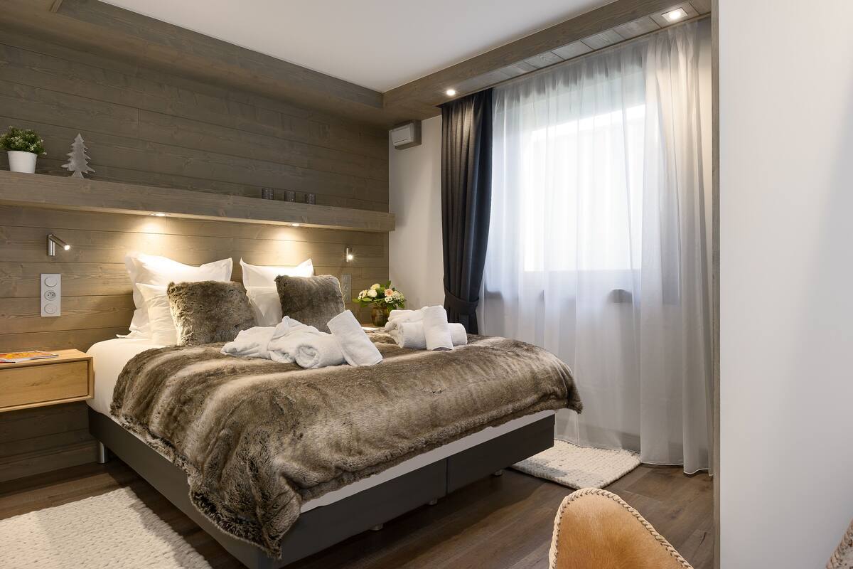A modern bedroom with a large bed, wooden headboard, bedside tables, and a window with sheer curtains.
