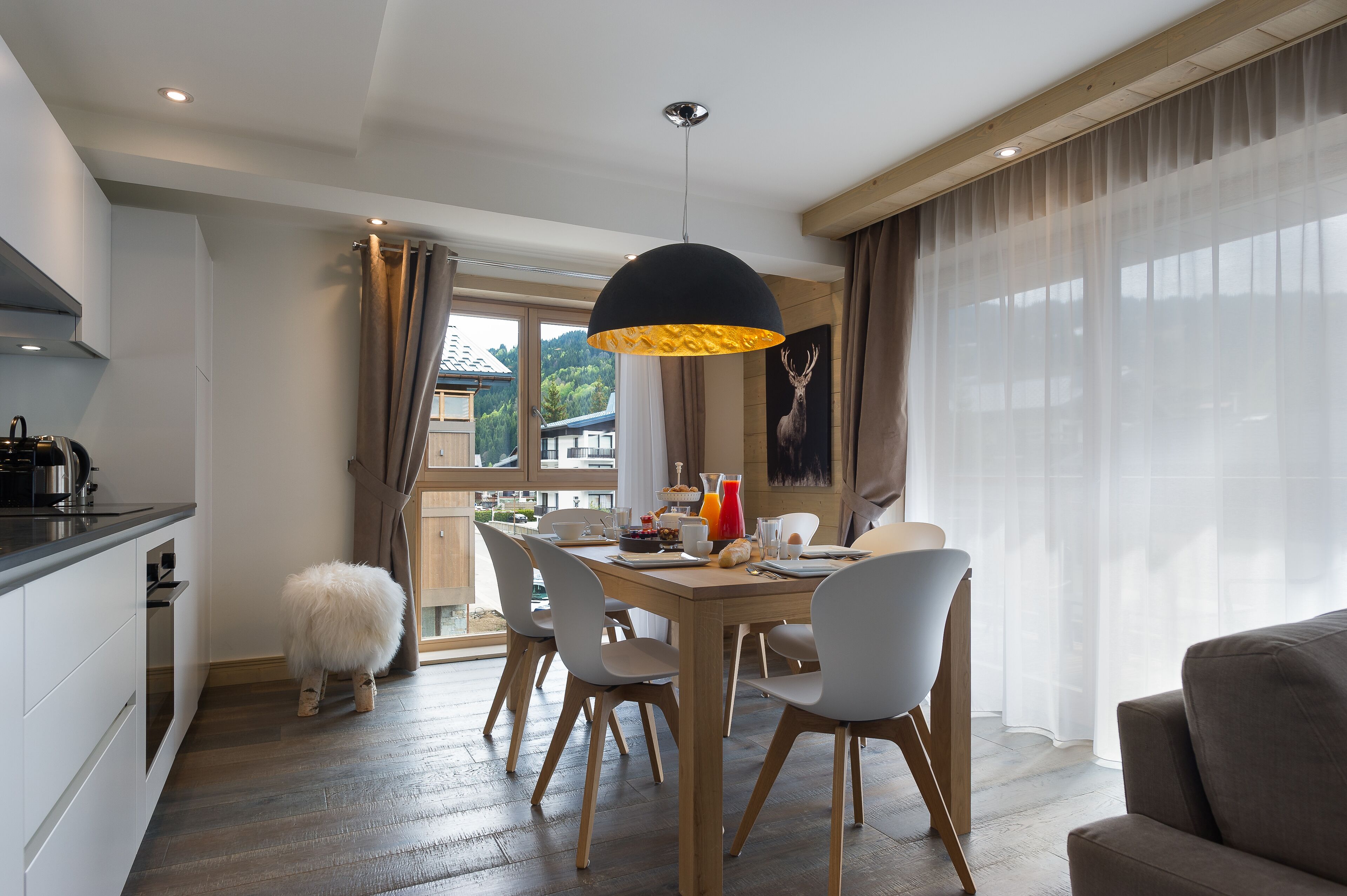 Un comedor con una mesa de madera, sillas blancas, un taburete mullido e una lámpara colgante negra.