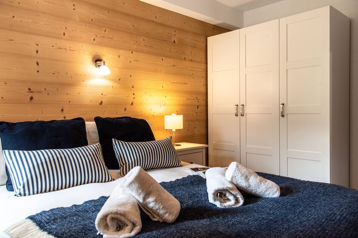 A hotel room with a wooden wall, a bed with striped pillows, a nightstand with a lamp, and a wardrobe.