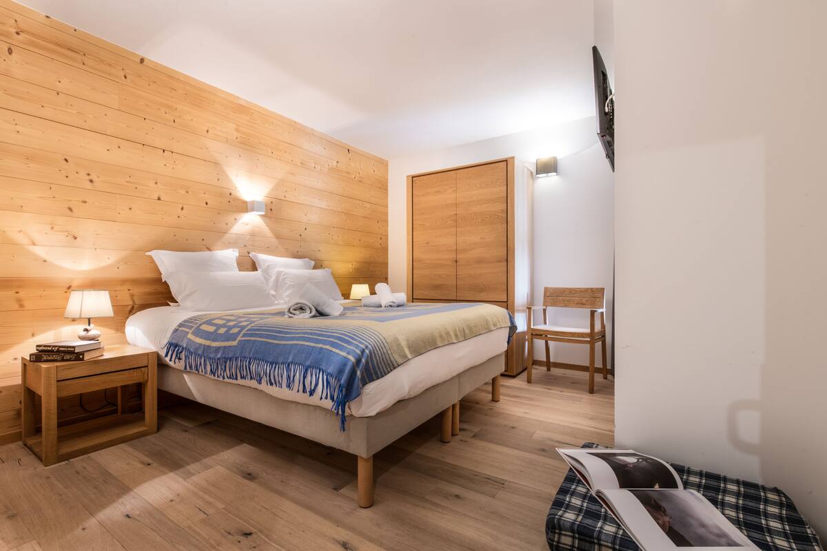 A neatly arranged bedroom with a wooden headboard, a bedside table with a lamp, a bed with a blue and white striped blanket, and a wooden wardrobe.