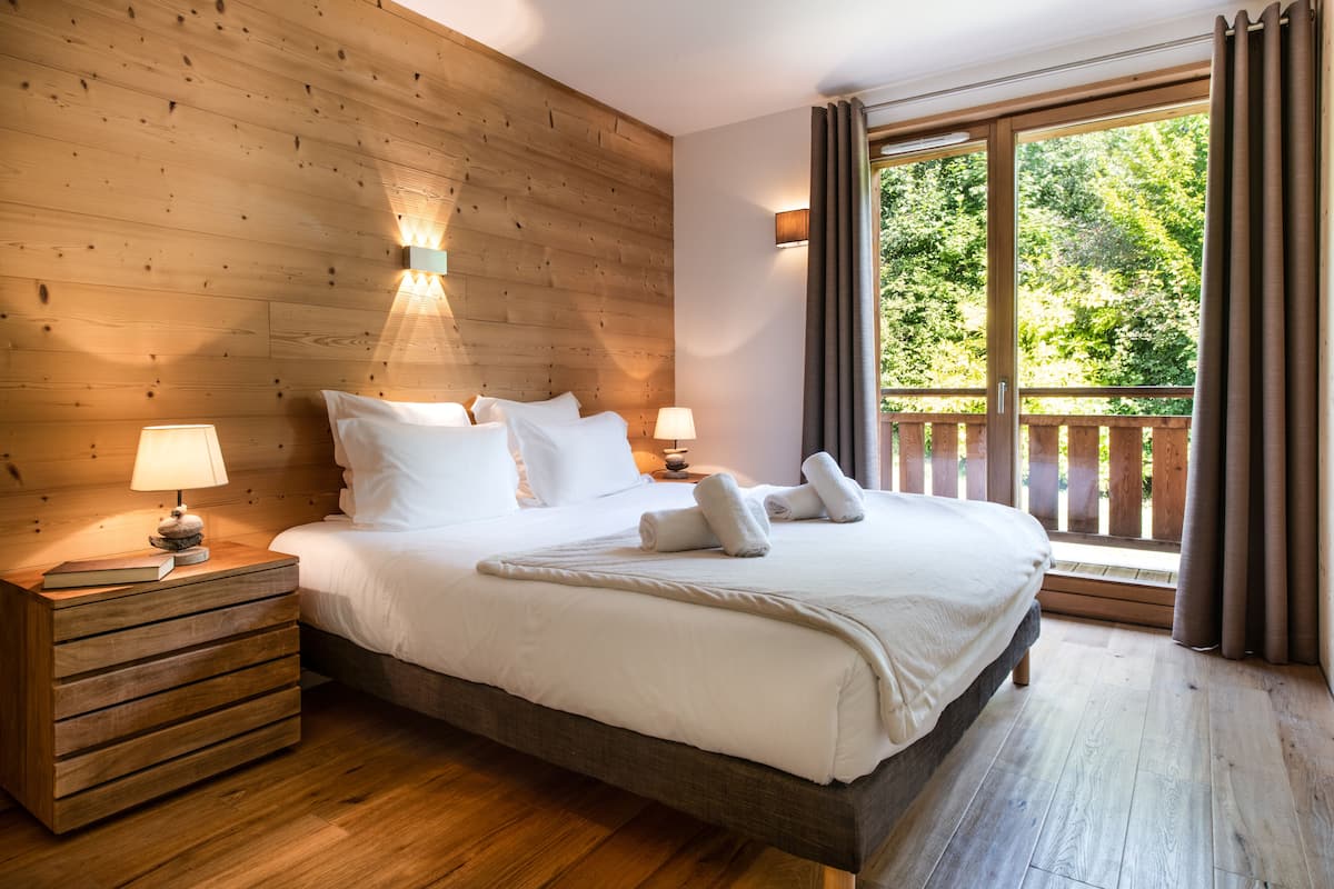 A wooden-paneled bedroom with a large bed, two bedside lamps, and a view of a balcony with curtains.