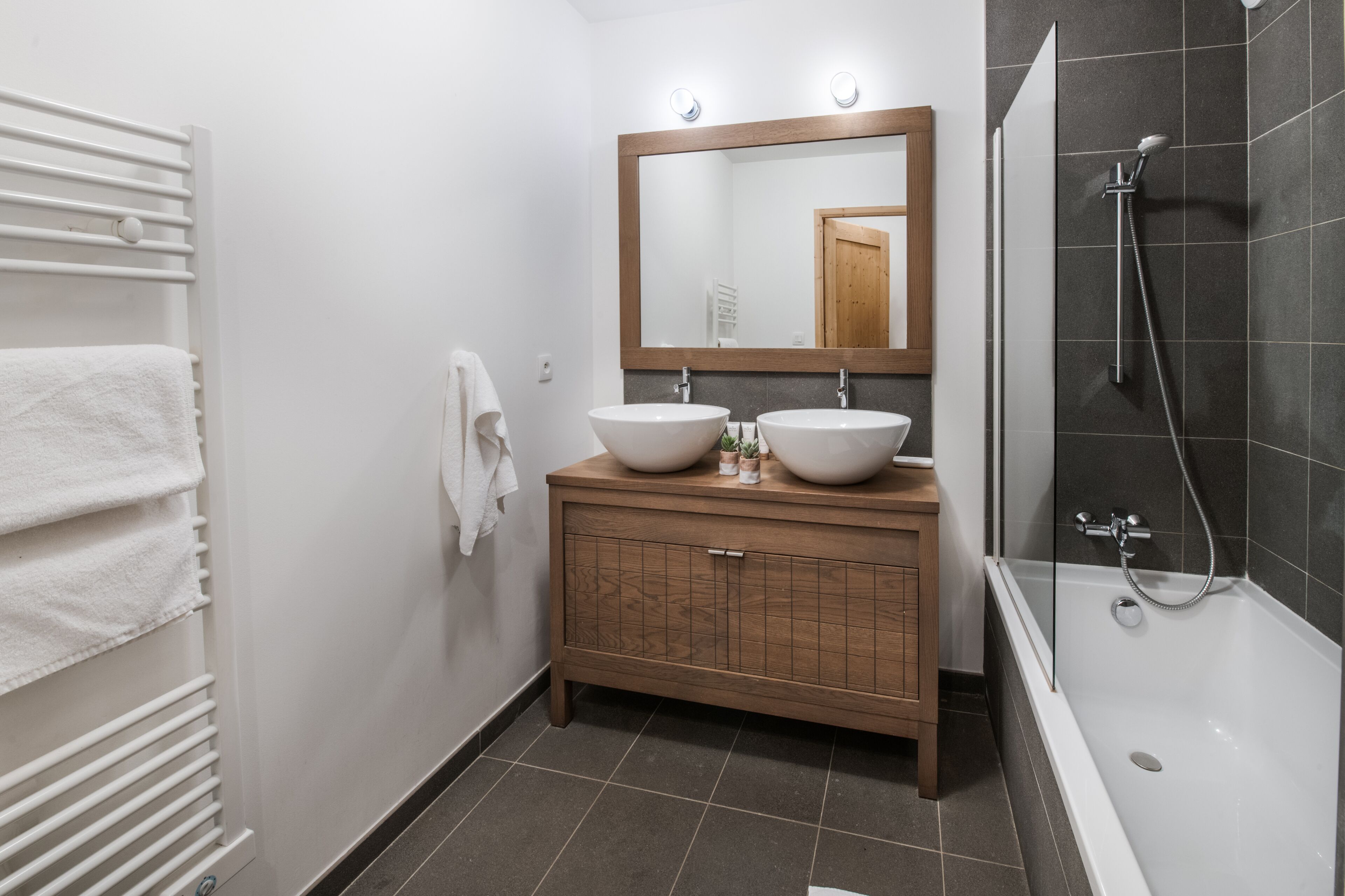 A modern bathroom with two sinks, a large mirror, a shower, and a wooden vanity.